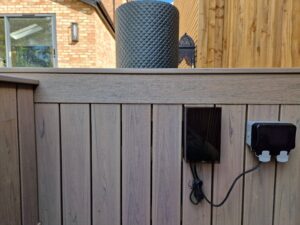 Picture shows an Inlite Hub and Hub Cover mounted on the fascia of a Trex Composite Deck beside an exterior plug socket